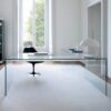 A minimalist transparent glass desk. The room has light-coloured walls and flooring, with a black chair behind the desk.
